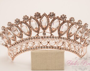Puede incluir: Una tiara de oro rosa con adornos de cristal transparente. La tiara está sobre un soporte beige. "Joli's Bridal" está escrito en rosa sobre fondo blanco debajo de la tiara.
