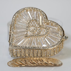 Pode incluir: Uma caixa de bugigangas dourada em forma de coração com um design transparente semelhante a cristal na tampa. A caixa está aberta, revelando um interior decorativo. Uma pequena pilha de moedas de ouro está abaixo. O logotipo "Yola's Bridal" está no canto inferior esquerdo.