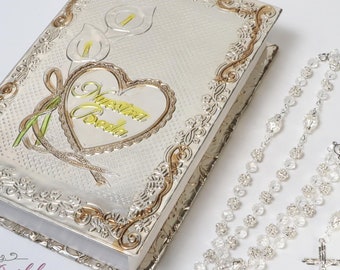 Puede incluir: Un libro plateado con diseños florales y de corazón, con la inscripción dorada "Nuestra Boda". Una rosario de cuentas transparentes con una cruz plateada está al lado. Para una boda o bautizo.