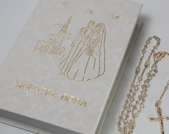Puede incluir: Libro de bodas de color marfil con ilustración dorada de una pareja y las palabras "Nuestra Boda". Un rosario con cuentas transparentes y una cruz dorada está al lado del libro. El libro tiene un patrón texturizado.