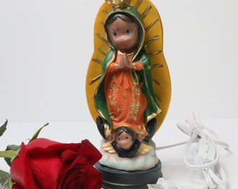 Puede incluir: Una figurita blanca, eléctrica, iluminada de la Virgen María, Nuestra Señora de Guadalupe, con una corona y un halo dorados. La figurita está de pie sobre una nube con un pequeño ángel a sus pies. Una rosa roja y un cable eléctrico blanco están en primer plano.