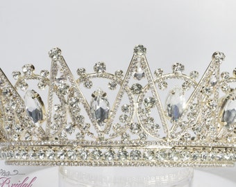 May include: A silver-toned bridal crown with clear, sparkling rhinestones. The crown features a series of pointed arches and large, teardrop-shaped stones. The base of the crown is lined with smaller rhinestones. The crown is displayed on a clear stand.