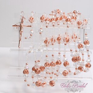 Può includere: Un set di rosari in cristallo e oro rosa esposti su un supporto in acrilico trasparente. I rosari sono realizzati con piccole perle di cristallo trasparente e perle più grandi in oro rosa. Il supporto ha quattro livelli ed è etichettato "Joli's Bridal Baptism and Special Occasion".