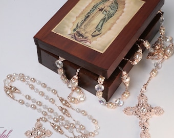 Puede incluir: Una caja de madera con una imagen de la Virgen María en la tapa. La caja contiene dos rosarios con cuentas de perla y cristal y cruces de oro rosa.