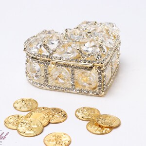 Op de afbeelding: Een hartvormige gouden doos met heldere kristallen versieringen en een deksel. De doos is open en er liggen gouden munten omheen.