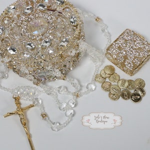 May include: A rosary with a gold crucifix, clear beads, and a round, jeweled container. A small, square, jeweled box and gold-colored religious medals are also present. The image includes a label that reads "Yeli's Mom Boutique."