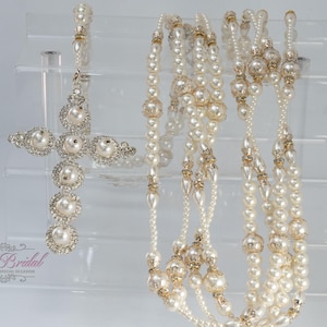 Puede incluir: Un rosario con un colgante de cruz adornado con perlas blancas y cristales transparentes, unido a una larga cadena de perlas blancas alternadas con cuentas de metal dorado. El texto Yoli's Bridal Baptism and Special Occasion es visible.