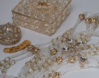 Puede incluir: Un surtido de accesorios de novia, que incluye una caja dorada con incrustaciones de cristal, un recipiente redondo de cristal y un collar largo de múltiples hilos con un colgante de cruz de cristal. También hay monedas de oro y cuentas decorativas.