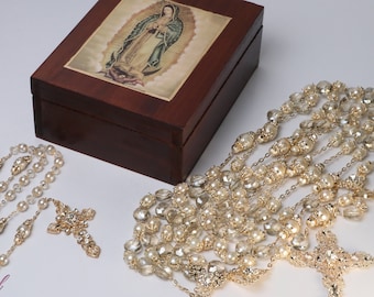 Puede incluir: Dos rosarios de perlas doradas y blancas con una caja de madera que presenta una imagen de la Virgen María. La caja está cerrada.