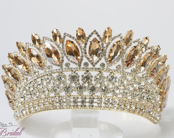 Puede incluir: Una tiara dorada con diamantes de imitación transparentes y ámbar. La tiara tiene una corona puntiaguda y una banda que se envuelve alrededor de la cabeza.