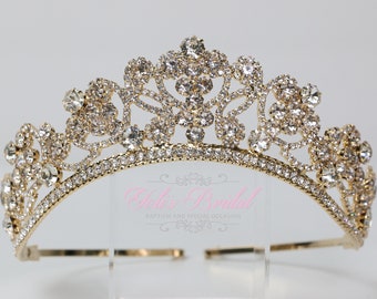 May include: A gold tiara with clear rhinestones, featuring a floral design. The tiara is on a clear stand.
