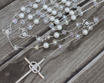 Puede incluir: Un rosario de cristal y perlas blancas y plateadas con una cruz plateada. El rosario está colocado sobre una superficie de madera.