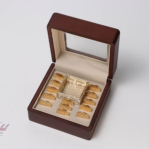 Op de afbeelding: Een donkerhouten doos met een doorzichtig deksel, gevoerd met crèmekleurige stof, bevat een goudkleurige decoratieve tray en diverse goudkleurige items. De doos is open en toont de inhoud. De tekst "Toli's Bridal" is zichtbaar.