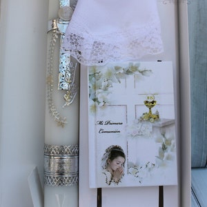 May include: A white box containing a white communion candle with silver trim, a white lace handkerchief, a small silver rosary, and a white card with the text "Mi Primera Comuni&oacute;n" and a picture of a girl praying.