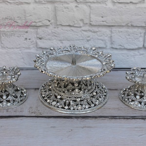 May include: A silver cake stand with two smaller matching stands. The stands are decorated with rhinestones and have a delicate, ornate design.
