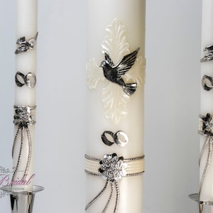 May include: Three white wedding unity candles with silver accents. The center candle features a dove and a cross design. The other two candles have infinity symbols and a silver bow.