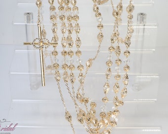 May include: Gold-coloured rosary with a cross pendant, displayed on a clear acrylic stand. The rosary features multiple strands of beads, with a mix of clear and gold-coloured elements. The cross is adorned with decorative accents.