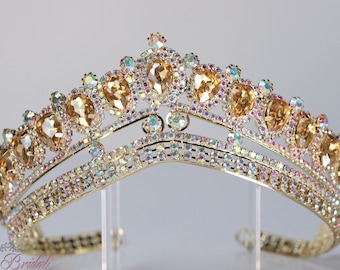 Puede incluir: Una tiara dorada con una doble fila de piedras de cristal iridiscentes y piedras grandes de color ámbar. La tiara está sobre un soporte transparente.