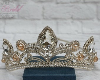 Puede incluir: Una tiara plateada y dorada con diamantes de imitación transparentes y melocotón. La tiara está sobre una caja azul.