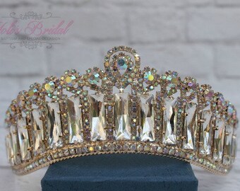 Puede incluir: Una tiara dorada con cristales transparentes e iridiscentes. La tiara tiene una pieza central puntiaguda y una forma curva.