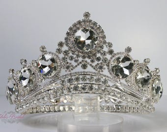 May include: A silver-toned tiara with large, clear, faceted gemstones and smaller, sparkling crystals. The crown features a central, prominent gemstone and a symmetrical design, suitable for special occasions.