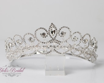 Könnte beinhalten: Eine silberne Tiara mit einem großen, klaren Kristall in der Mitte und kleineren Kristallen, die sie umgeben. Die Tiara hat ein zartes, wirbelndes Design.