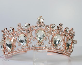 May include: A rose gold tiara with clear, teardrop-shaped and round crystals. The crown features a delicate, ornate design with multiple crystal accents. A bridal or special occasion headpiece.