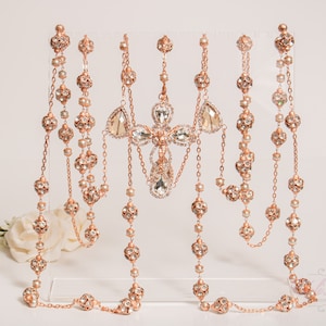 Può includere: Una collana a catena in oro rosa con un grande ciondolo a croce di cristallo e perline di cristallo più piccole. La collana è esposta su un supporto trasparente.