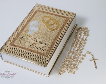 Puede incluir: Una caja dorada con interior blanco y un diseño decorativo con dos anillos, flores y el texto "Nuestra Boda". También se ve un rosario dorado con una cruz.