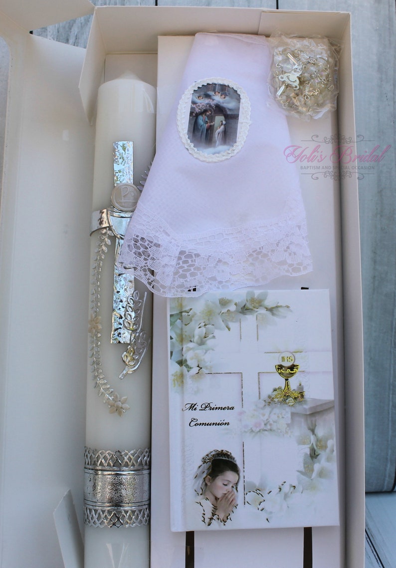 May include: A white box containing a white communion candle with silver trim, a white lace handkerchief, a small silver rosary, and a white card with the text "Mi Primera Comuni&oacute;n" and a picture of a girl praying.