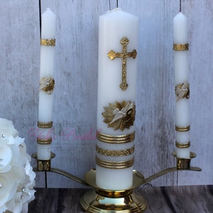 May include: A set of three white wedding unity candles with gold accents. The center candle features a gold cross and a pair of doves with a ring. The two smaller candles have gold rings and a gold design.