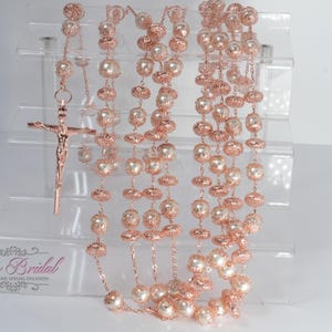 Può includere: Rosario in oro rosa con perline a forma di perla e dettagli decorativi in filigrana. È attaccato un crocifisso in oro rosa. Il rosario è esposto su un supporto in acrilico trasparente. Il logo "Yolis Bridal" è visibile.