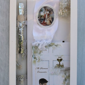 May include: A white box containing a white candle with silver accents, a white satin ribbon with a religious image, a white card with the text "Mi Primera Comuni&oacute;n" and a white card with the text "IHS".