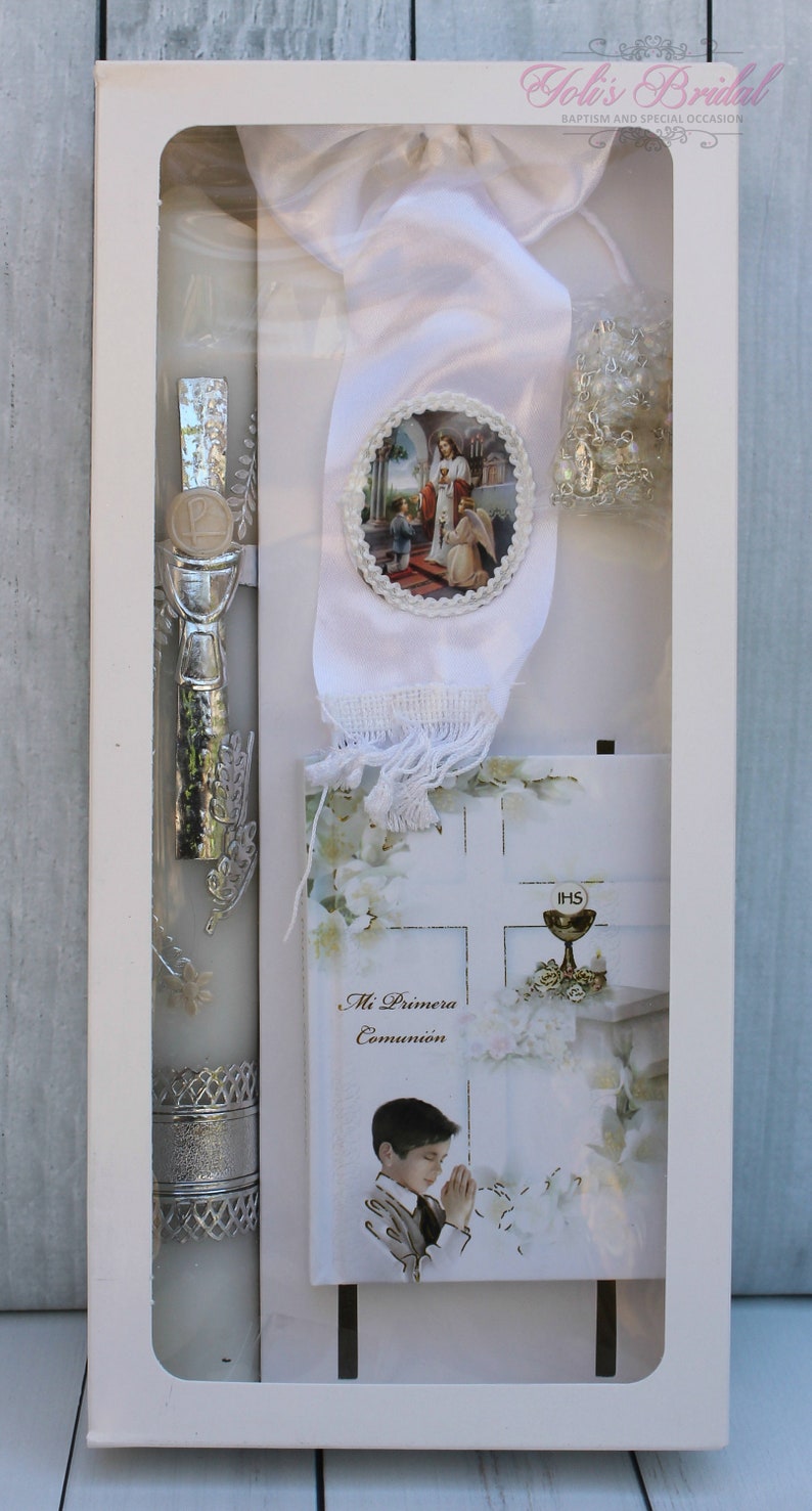 May include: A white box containing a white candle with silver accents, a white satin ribbon with a religious image, a white card with the text "Mi Primera Comuni&oacute;n" and a white card with the text "IHS".