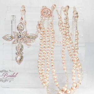 Può includere: Elegante set di gioielli da sposa con un pendente a croce e collane di perle a strati. Il pendente a croce è adornato con cristalli trasparenti e dettagli in oro rosa. Le collane hanno più fili di perle finte. I gioielli sono esposti su un supporto in acrilico trasparente.