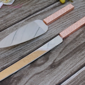 May include: A set of two silver cake knives with rose gold handles decorated with rhinestones. The handles are a sparkly rose gold color.