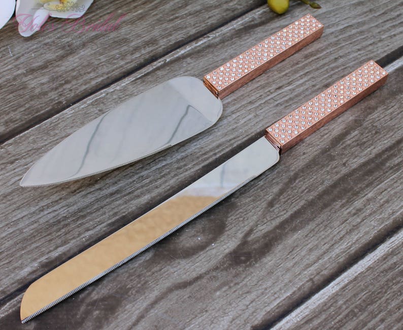 May include: A set of two silver cake knives with rose gold handles decorated with rhinestones. The handles are a sparkly rose gold color.