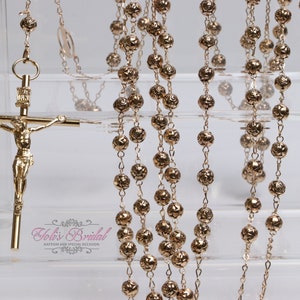May include: Gold-toned rosary with detailed beadwork and a crucifix. The rosary features a detailed figure of Jesus on the cross. The beads have a textured, floral-like design. The rosary is displayed on a clear, tiered surface.