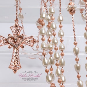 May include: A rose gold and white pearl rosary with a detailed rose gold cross. The cross has a heart-shaped design with clear rhinestones.