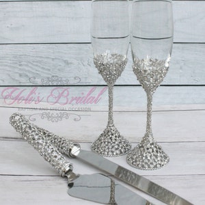 Può includere: Un set di due flute da champagne argentate con decorazioni in cristallo e un coltello da torta argentato con manico in cristallo. Le flute e il coltello sono su una superficie in legno bianco.