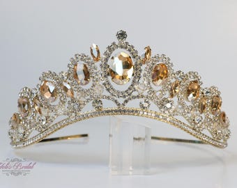 Puede incluir: Una tiara nupcial plateada adornada con cristales transparentes y ámbar. La tiara presenta un cristal central ámbar de forma ovalada, rodeado de piedras más pequeñas. El diseño incluye una base delicada y curvada y se exhibe sobre un soporte transparente.