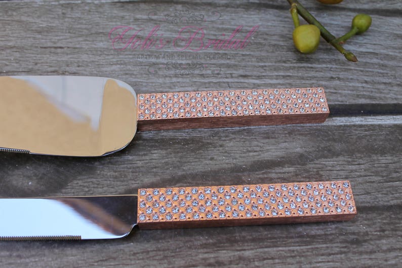 May include: A set of two cake cutting knives with rose gold handles and clear rhinestone embellishments.