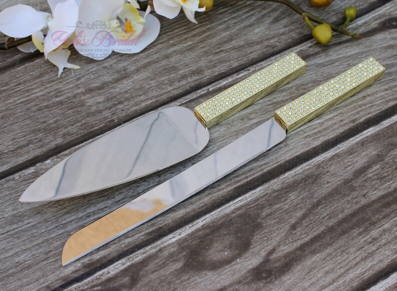 Puede incluir: Un juego de dos cuchillos para cortar pastel dorados con mangos de diamantes de imitaci&oacute;n transparentes. Los cuchillos est&aacute;n sobre una superficie de madera.