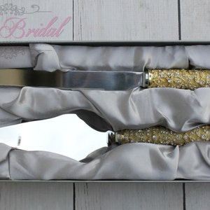 Könnte beinhalten: Ein Set goldener Kuchenschneidemesser mit Strasssteinen, in einer grau satinierten Box. Die Messer sind für eine Hochzeit oder einen besonderen Anlass.