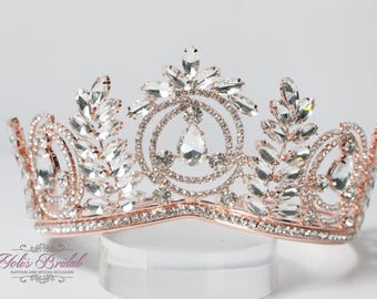 Puede incluir: Una tiara de oro rosa con adornos de cristal transparente. La tiara presenta un diseño central de dos círculos superpuestos con un cristal en forma de lágrima en el centro. La tiara está decorada con cristales en forma de hoja y cristales redondos más pequeños.