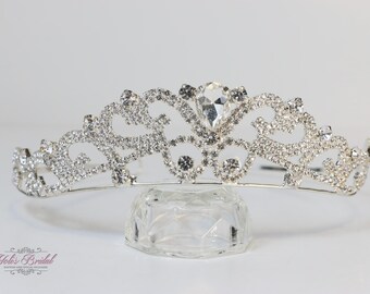 Könnte beinhalten: Eine silberne Tiara mit klaren Strasssteinen und einem großen, tropfenförmigen Mittelstein. Die Tiara wird auf einem klaren, facettierten Ständer präsentiert, ideal für Hochzeiten oder besondere Anlässe.