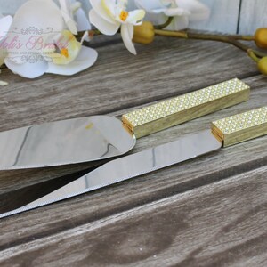 Puede incluir: Un juego de dos cuchillos para cortar pastel dorados con mangos de diamantes de imitaci&oacute;n transparentes. Los cuchillos est&aacute;n sobre una superficie de madera.