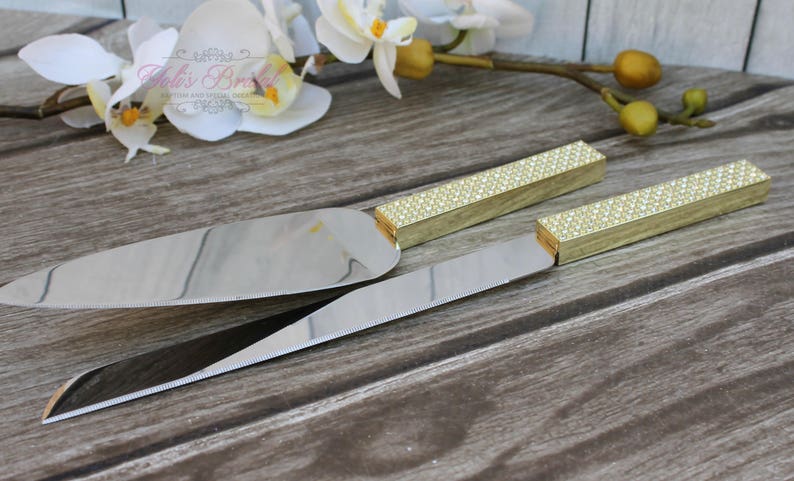 Puede incluir: Un juego de dos cuchillos para cortar pastel dorados con mangos de diamantes de imitaci&oacute;n transparentes. Los cuchillos est&aacute;n sobre una superficie de madera.