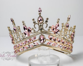 Puede incluir: Una corona dorada con diamantes de imitación rosas y transparentes. La corona tiene una parte superior puntiaguda y una base ancha.