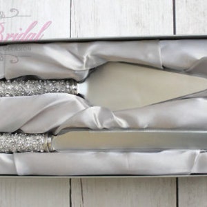 Könnte beinhalten: Ein silbernes Kuchenschaber- und -Server-Set mit Strasssteinen in einer weiß satinierten Box. Das Set ist zum Schneiden und Servieren von Hochzeitstorte.
